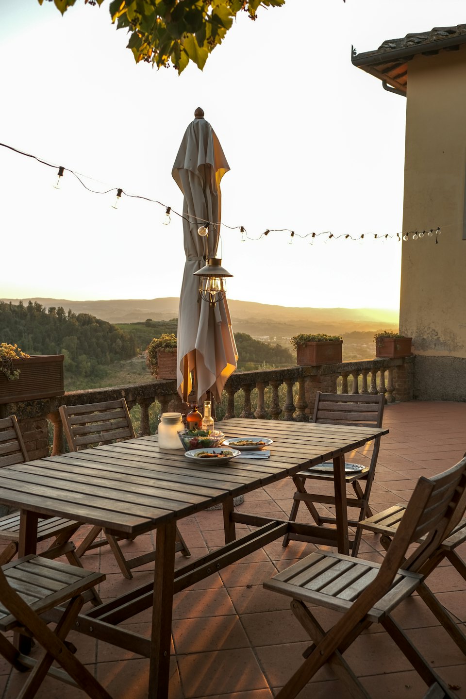 Outdoor terrace dining table with food and drinks overlooking sunset hills.