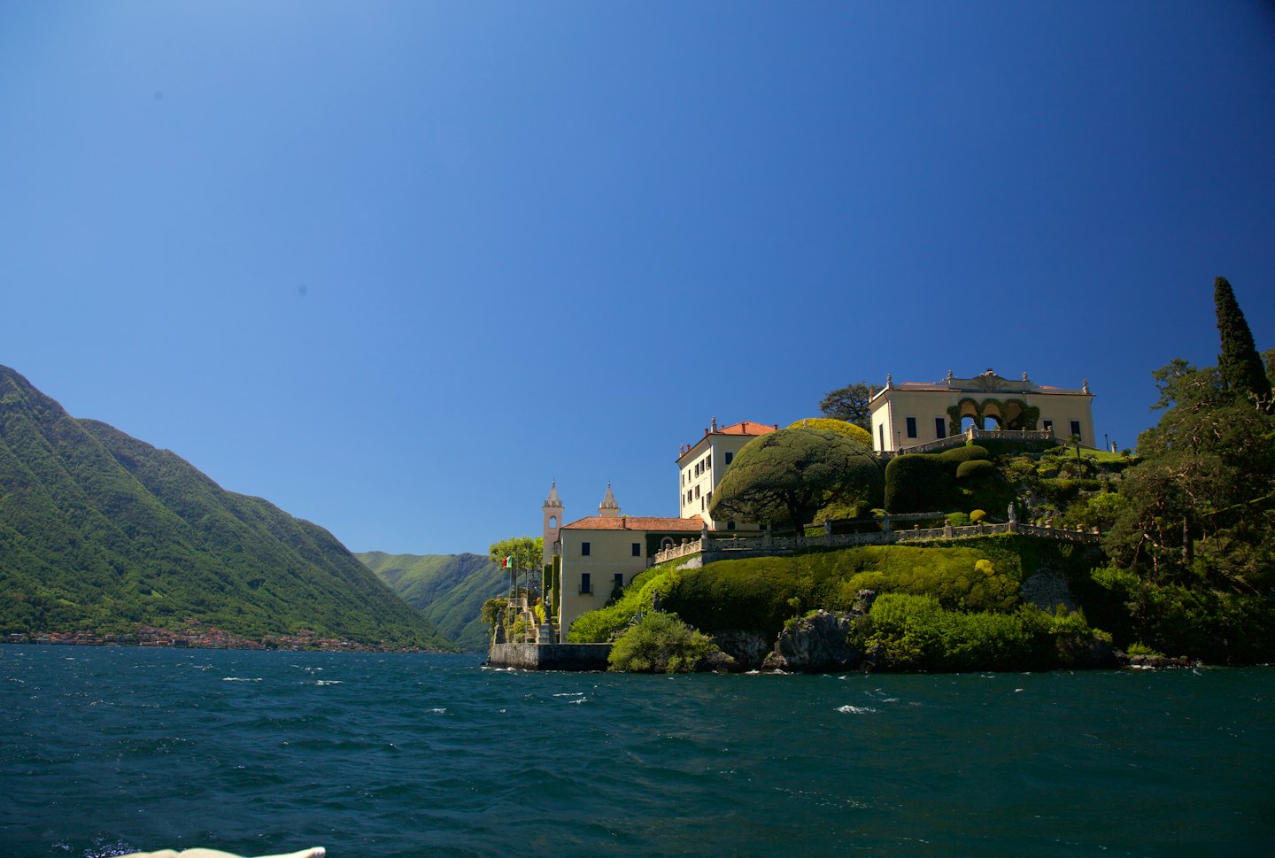 A large villa sits on a green hillside by a lake.